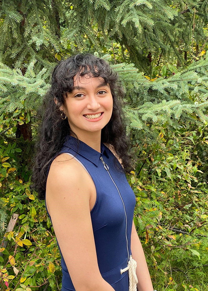 BLaST Scholar Daphne Mueller is smiling and standing in front of green spruce trees and foliage outside in the sun.