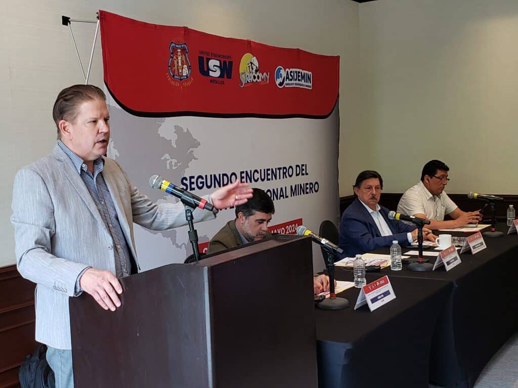 An image of a person standing and speaking behind a podium. Beside the podium is a table with three people sitting behind it. 