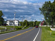 A maroon car traveling away from the viewer on a paved road with two yellow lines down the middle, one solid and the other dashed, and white lines on the sides as it goes over a small rise between a white two-story wooden house on the left and a red gambrel-roofed barn on the right. White wooden fences are along both sides of the road and a small blank yellow reflectorized marker sits on a metal post at the right. Above the sky is streaked with cloud; sunlight illuminates the scene from the left