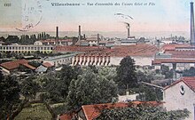 Ancienne carte postale colorisée. Vue en plongée sur des usines desquelles dépassent cinq longues cheminées