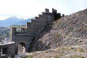 Crenellated stone wall following the contour of a rock with a door, at the top of which there is a dish accessible from the right.