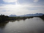 A wide flat river flows between tree-lined banks. Jagged hills rise in the distance.