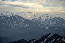 Montañas boscosas abajo y montañas nevadas arriba, bajo un cielo nublado.