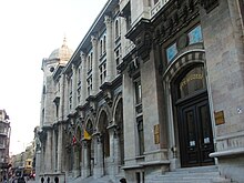 An arched neoclassical building with hanging banners, with a yellow vehicle parked in front