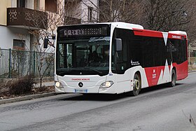 Photographie en couleurs d’un autobus de marque Mercedes-Benz rouge et blanc dans un environnement urbain et descendant une rue en légère pente.