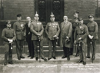 Photo noir et blanc de neuf hommes debout, dans une rue, devant la porte d'entrée fermée d'un bâtiment. Sept hommes sont en tenue militaire. Il s'agit d'Adolf Hitler et des autres personnalités inculpées lors du son procès en 1924.