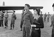 Photo en noir et blanc. Des hommes en uniforme et une femme en tailleur noir (la reine) observent le ciel, sur un terrain herbeux.