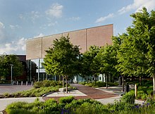 Photograph of the exterior entrance to the Wichita Art Museum in May 2020 showing the brick and concrete Lattner and Walker Family Plaza and the doors and floor-to-ceiling windows of the museum's main entrance, which is surrounded by trees and landscaping