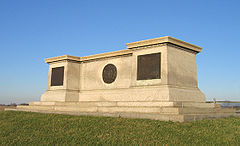 A large monument of off-white stone. Its shape resembles a long, flat pedestal. It sits atop a grassy knoll with a bright blue sky overhead.