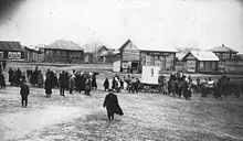 Group of people in a clearing, with wooden buildings in the background