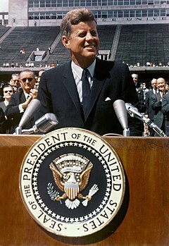 Kennedy đứng phát biểu trước bục gỗ tại Đại học Rice. Phó Tổng thống Lyndon Johnson và các quan chức khác đứng ở phía sau.