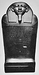 Black and white image of a dark stone coffin with a human face, the coffin stands upright facing the viewer.