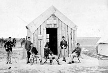 Photo en noir et blanc d'un groupe d'hommes en uniforme devant un bâtiment en bois.