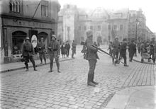 Černobílá fotografie s ozbrojenými vojáky v helmách v ulicích města, v pozadí jsou přihlížející lidé