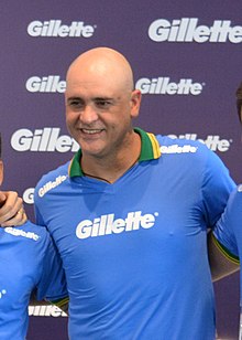 Um homem branco, careca, sorrindo e não olhando pra câmera. Ele veste uma camisa azul escrito "Gillette".