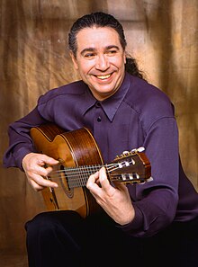 Laucke, seated in half profile, playing his guitar and smiling while looking away from the camera