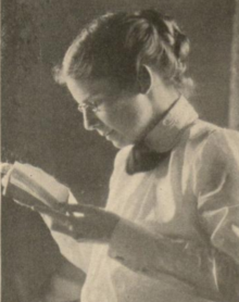 A young white woman, hair in a bun, wearing a high-collared white blouse, holding and regarding a small book or pamphlet