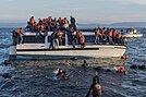 Syrian and Iraqi immigrants getting off a boat from Turkey on the Greek island of Lesbos. A small boat in water, with land on the horizon behind. Many people are on its outside in orange life jackets, some carrying inner tubes as well. A few are in the water swimming toward the camera. In the foreground a man in a red and black wetsuit has his hand out to them.