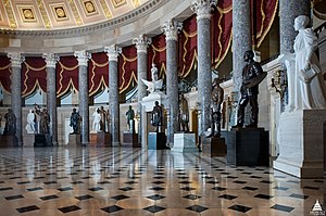 A line of statues along the wall of a circular room. Statues are interspersed between columns. Red curtains are situated behind the statues. A large dome is visible above, enclosing the room.