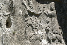 photo d'un mur sculpté, on voit deux hommes face à face tenant chacun une coupe, aux pieds du deuxième, un chien