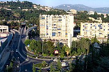 Vue d'un immeuble en ville contourné par une bretelle de voie rapide menant vers un tunnel sous une colline boisée.