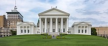 An all white Neoclassical building with pediment and six columns rises on a grassy hill with a large American elm tree in the left foreground. Two boxier, but similarly styled wings are attached at the building's rear.