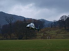 A helicopter is seen landing in a spots field. Goal posts and a small hill are seen at the end of the field behind the helicopter, and in the far distance larger hills or mountains