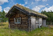 Dřevěný domek se střechou porostlou trávou, vzadu a kolem stromy