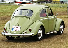 Picture of a yellow automobile in a field, with an airplane in the background. This is a rear three-quarters-view of it. It has a beetle-like body shape, and its wheels are compact-sized.