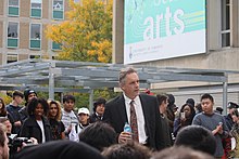 Jordan Peterson con una botella de agua en la mano en medio de una multitud congregada al frente de un edificio en un día soleado.