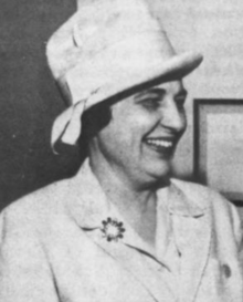 A smiling white woman with dark hair, wearing a white brimmed hat with a high crown, and a white suit