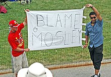 Photo de deux hommes tenant une bannière blanche sur laquelle est écrit "Blame Mosley"