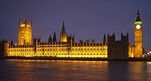 Large sand-coloured building of Gothic design beside brown river and road bridge. The building has several large towers, including large clock-tower.