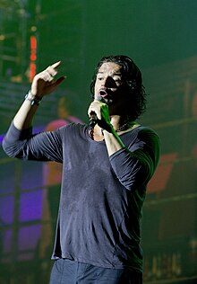 A man with blue jeans and shirts half-opened holding a microphone with his right hand while singing in front of a stage, with a city simulated background behind him.