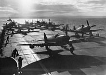 The aft section of an aircraft carrier's flight deck. Several propeller aircraft are parked here, and are being worked on.