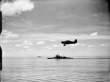 Photo en noir et blanc d'un avion à hélices survolant un navire de guerre