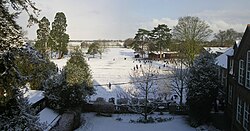 A panoramic vista of school playing fields covered in snow. Two large trees are visible to the left, schoolrooms to the right. A number of pupils are seen playing in the snow.