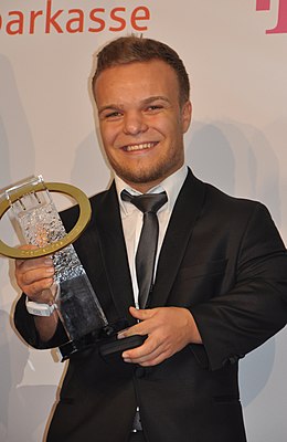 Kappel hält einen gläsernen mit einem goldenen Ring ausgestatteten Preis. Er trägt einen schwarzen Anzug und ein helles Hemd mit schwarzer Krawatte. Sein Haar ist kurz und blond.