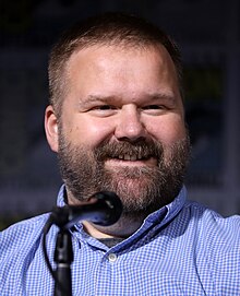 A man with a beard, smiling and wearing a blue checkered shirt, speaks into a microphone at a panel.