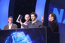 Four men stand behind a podium, one speaking into a microphone with his arms outstretched, holding an award.