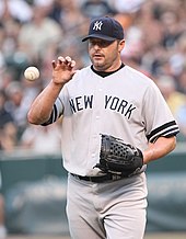 A close-up view of a man on a pitcher's mound, wearing a Yankees uniform. He has a glove on his left hand, positioning to catch a mid-air baseball bare-handed with his right.