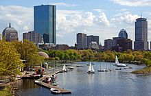 Skyline of a city on a river. Two buildings are much taller and more prominent than the rest; one of the large buildings is a skyscraper with a blue-tinted, all-glass facade, and the second is a large rectangular tower with a steel latticework facade and a tall antenna mast on its roof.