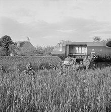 Photo en noir et blanc de soldats en tenue de combat près d'un planeur enfoncé dans un muret. Ils sont dissimulés par les cultures des champs à proximité. En arrière-plan sont visibles une maison et quelques arbres.