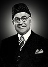 A black-and-white head and shoulder shot of a man with spectacles, wearing coat and a tie.