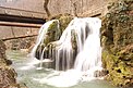 Über einen Travertin-Felsen stürzt das Wasser breit gestreut mehrere Meter in die Tiefe. Am oberen Rand sieht man im Hintergrund zwei Fußgängerbrücken.