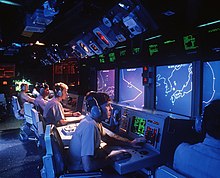 A row of monitors with maps, charts and data with operators sitting in front