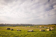 Foto einer Hügelkuppe mit in einem Kreis angeordneten Steinen und drei Bänken. Im Hintergrund sieht man städtische Bebauung.