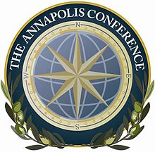 Un logo circulaire, semblable à un sceau. Au centre une rose des vents jaune superposée à un globe stylisé bleu. Autour, une couronne jaune avec les quatre points cardinaux. Autour, une bande bleue cerclée de jaune avec écrit The Annapolis Conference. En bas, des rameaux d’olivier épousent la forme du sceau.