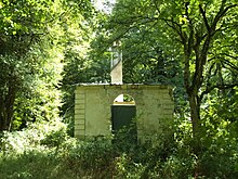 Photographie en couleurs d'une maisonnette ruinée envahie par la végétation d'un sous-bois.