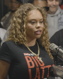 Latto wearing a black "BIG LATTO" t-shirt and gold chain, at the mic with a crowd in back.